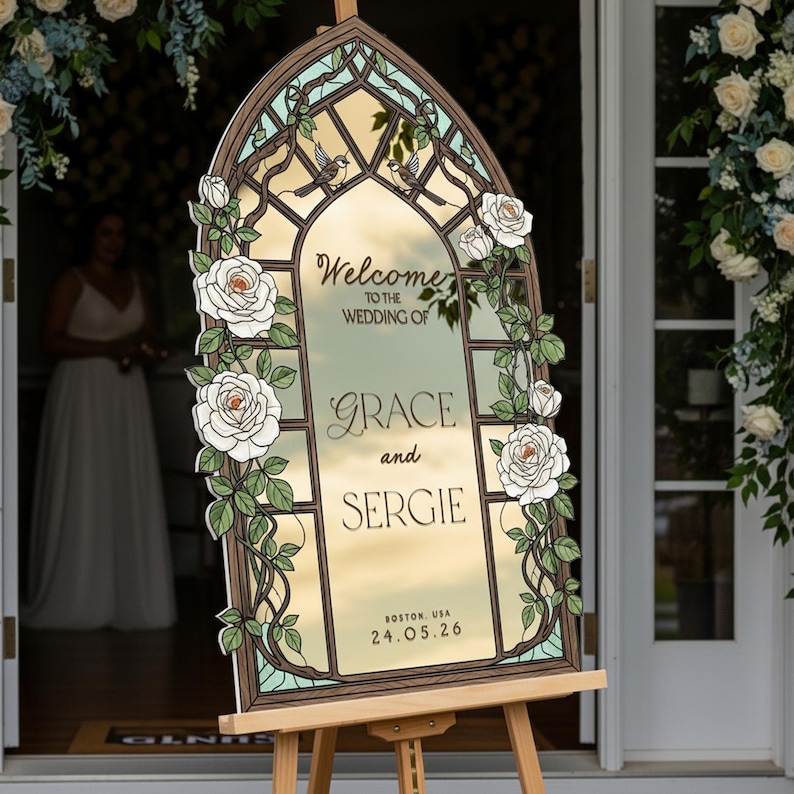 May include: An arched wedding welcome sign featuring a stained glass window design with white roses, green vines, and two small birds. The text reads: Welcome to the wedding of Grace and Sergie, Boston, USA, 24.05.26. The sign is displayed on a wooden easel.