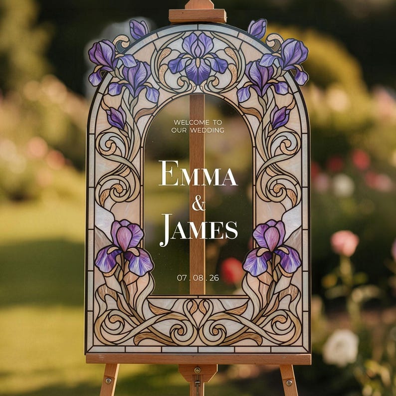 May include: An arched wedding sign featuring a stained glass style border with purple iris flowers and tan scrollwork. The center text reads: WELCOME TO OUR WEDDING, EMMA & JAMES, 07.08.26. The sign is mounted on a wooden easel frame.
