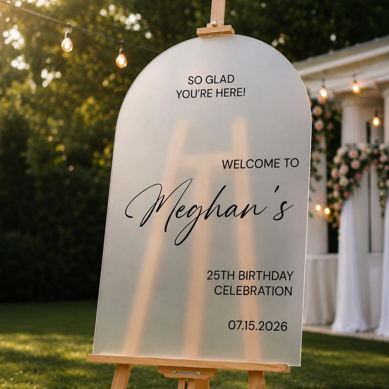 May include: An arched, frosted acrylic welcome sign on a wooden easel. The text reads: SO GLAD YOU'RE HERE! WELCOME TO Meghan's 25TH BIRTHDAY CELEBRATION 07.15.2026. The sign features a semi-transparent surface with black printed lettering.