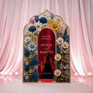 May include: An arched wedding welcome sign with a deep red center panel featuring the white text WELCOME TO THE WEDDING OF OLIVIA and MARCUS JUNE 24, 2026. The frame is decorated with a colorful floral border of blue, yellow, and white flowers.