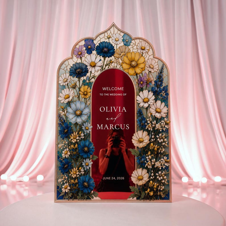 May include: An arched wedding welcome sign with a deep red center panel featuring the white text WELCOME TO THE WEDDING OF OLIVIA and MARCUS JUNE 24, 2026. The frame is decorated with a colorful floral border of blue, yellow, and white flowers.