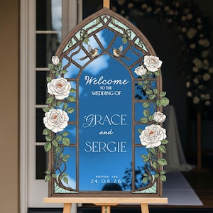 May include: An arched wedding welcome sign featuring a stained glass window design with white roses, green leaves, and two small birds. The blue center panel displays the text: Welcome to the wedding of Grace and Sergie, Boston, USA, 24.05.26.