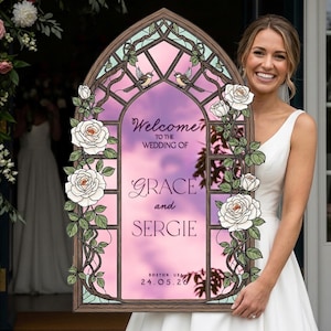 May include: An arched wedding welcome sign featuring a stained glass window design with white roses, green vines, and two small birds. The center displays the text: Welcome to the wedding of Grace and Sergie, Boston, USA, 24.05.26, over a pink sky.