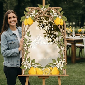 May include: A wooden framed sign with a mirror center, decorated with lemons, leaves, and white flowers. The sign reads "Welcome to AMANDA'S Bridal Shower" and "March 25, 2026". The sign is on an easel.