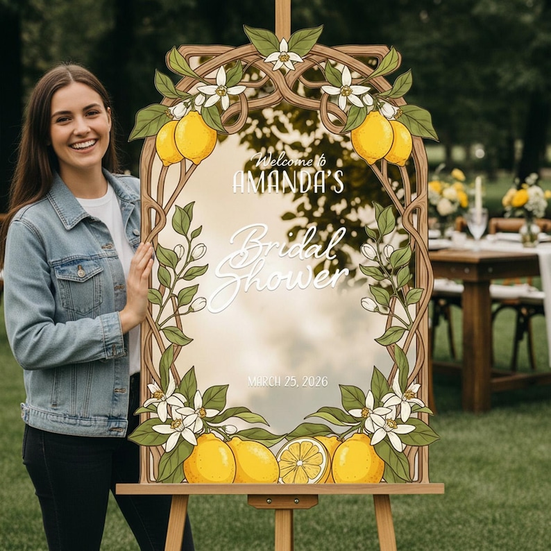 May include: A wooden framed sign with a mirror center, decorated with lemons, leaves, and white flowers. The sign reads "Welcome to AMANDA'S Bridal Shower" and "March 25, 2026". The sign is on an easel.