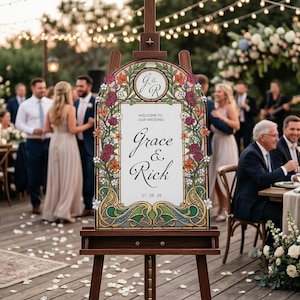 K&ouml;nnte beinhalten: Eine h&ouml;lzerne Staffelei mit einem Hochzeits-Willkommensschild. Das Schild zeigt die Namen "Grace & Rick" mit floralen und Jugendstil-Bord&uuml;ren. Das Datum "07.08.26" wird ebenfalls angezeigt. Der Hintergrund zeigt eine Hochzeitsfeier.