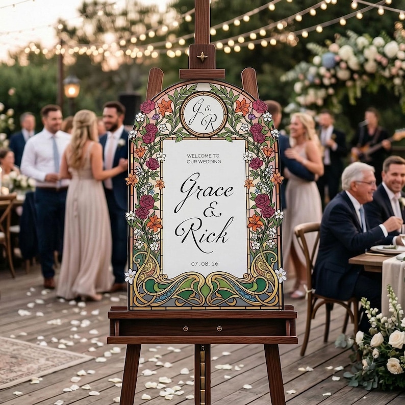 K&ouml;nnte beinhalten: Eine h&ouml;lzerne Staffelei mit einem Hochzeits-Willkommensschild. Das Schild zeigt die Namen "Grace & Rick" mit floralen und Jugendstil-Bord&uuml;ren. Das Datum "07.08.26" wird ebenfalls angezeigt. Der Hintergrund zeigt eine Hochzeitsfeier.