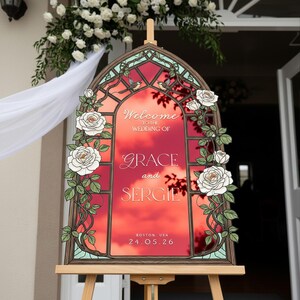 May include: An arched wedding welcome sign featuring a stained glass window design with white roses and green vines. The center displays the text: Welcome to the wedding of Grace and Sergie, Boston, USA, 24.05.26, set against a vibrant red sunset background.