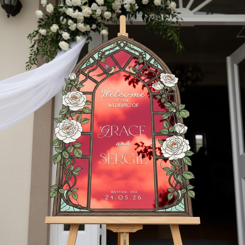 May include: An arched wedding welcome sign featuring a stained glass window design with white roses and green vines. The center displays the text: Welcome to the wedding of Grace and Sergie, Boston, USA, 24.05.26, set against a vibrant red sunset background.