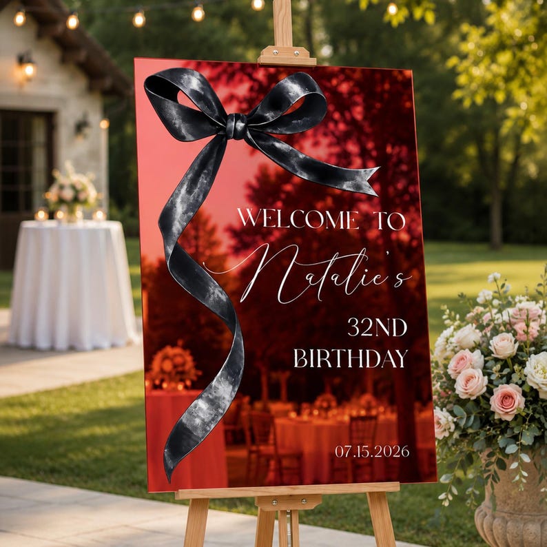 May include: A rectangular red acrylic welcome sign on a wooden easel featuring a large black ribbon graphic. The white text reads: WELCOME TO Natalie's 32ND BIRTHDAY 07.15.2026. The surface is reflective, showing a blurred outdoor event setting.