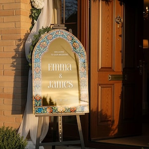 May include: An arched gold mirror wedding sign featuring a colourful stained glass-style floral border. The text reads: WELCOME TO OUR WEDDING, EMMA & JAMES, 05.18.28. The sign is displayed on a black metal easel stand in front of a wooden door.