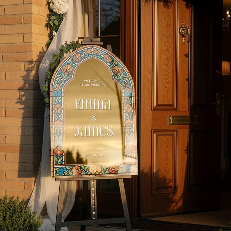 May include: An arched gold mirror wedding sign featuring a colourful stained glass-style floral border. The text reads: WELCOME TO OUR WEDDING, EMMA & JAMES, 05.18.28. The sign is displayed on a black metal easel stand in front of a wooden door.