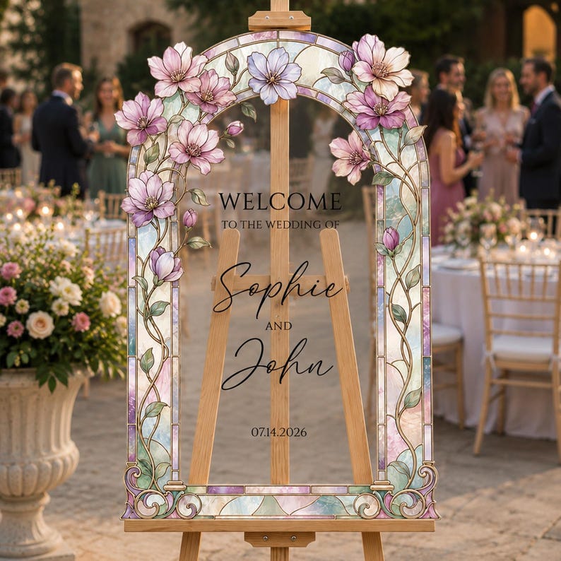 May include: A wedding welcome sign on a wooden easel featuring a stained glass arch frame decorated with pink magnolia flowers and vines. The text reads: WELCOME TO THE WEDDING OF Sophie AND John 14.07.2026 in black lettering on a clear panel.