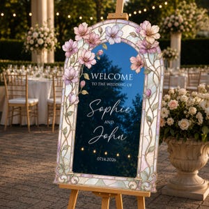 May include: An arched wedding welcome sign on a wooden easel featuring a stained glass border with pink and purple floral illustrations. The dark blue centre displays the text: Welcome to the wedding of Sophie and John, dated 14.07.2026.