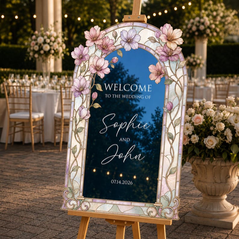 May include: An arched wedding welcome sign on a wooden easel featuring a stained glass border with pink and purple floral illustrations. The dark blue centre displays the text: Welcome to the wedding of Sophie and John, dated 14.07.2026.