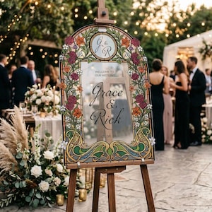 K&ouml;nnte beinhalten: Eine h&ouml;lzerne Staffelei zeigt ein dekoratives Hochzeits-Willkommensschild. Das Schild hat einen floralen Rand mit burgunderroten, orangefarbenen und wei&szlig;en Blumen. Der Text lautet "Willkommen zu unserer Hochzeit" und die Namen "Grace & Rich" mit dem Datum 07.08.26.