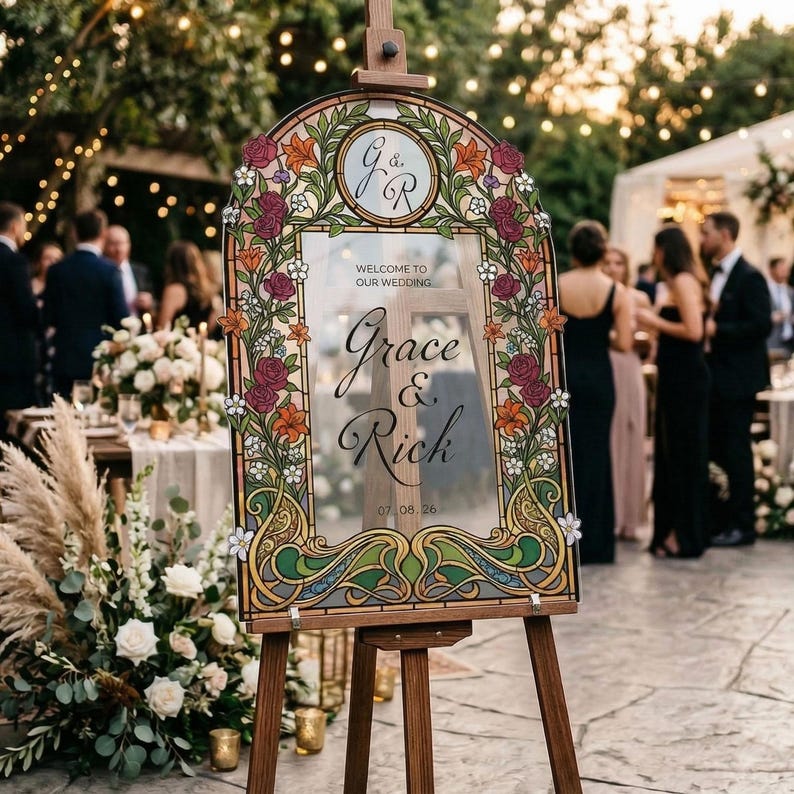 K&ouml;nnte beinhalten: Eine h&ouml;lzerne Staffelei zeigt ein dekoratives Hochzeits-Willkommensschild. Das Schild hat einen floralen Rand mit burgunderroten, orangefarbenen und wei&szlig;en Blumen. Der Text lautet "Willkommen zu unserer Hochzeit" und die Namen "Grace & Rich" mit dem Datum 07.08.26.