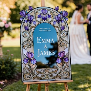 May include: An arched wedding welcome sign featuring a stained glass-style border with purple iris flowers and gold scrollwork. The center is a blue panel with the white text: WELCOME TO OUR WEDDING, EMMA & JAMES, 07.08.26, displayed on a wooden easel.