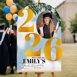 May include: A graduation party sign on an easel. The sign features the text "Class of 2026" and "Welcome to Emily's Graduation Party". A photo of a graduate is in the center. Gold numbers and text are on a blue background.