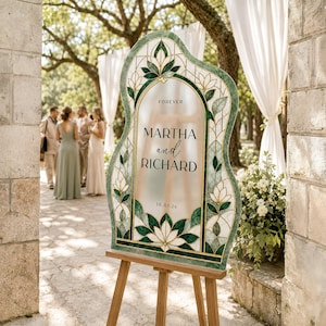 May include: An arched wedding sign with a green and white stained glass-style border featuring floral motifs. The center is frosted with the text FOREVER MARTHA and RICHARD 10.01.26 in black lettering. The sign is displayed on a wooden easel.