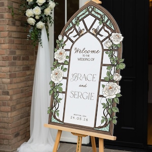 May include: An arched wedding welcome sign featuring a stained glass window design with white roses, green vines, and two birds. The text reads: Welcome to the wedding of Grace and Sergie, Boston, USA, 24.05.26. The sign is displayed on a wooden easel.