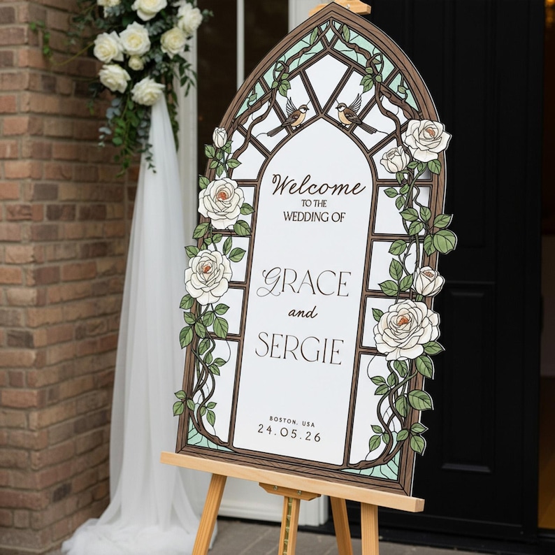 May include: An arched wedding welcome sign featuring a stained glass window design with white roses, green vines, and two birds. The text reads: Welcome to the wedding of Grace and Sergie, Boston, USA, 24.05.26. The sign is displayed on a wooden easel.