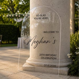 May include: An arched clear acrylic sign featuring black text that reads: SO GLAD YOU'RE HERE! WELCOME TO Meghan's 25TH BIRTHDAY CELEBRATION 07.15.2026. The sign has a smooth, transparent surface and a rounded top edge, displaying custom lettering.