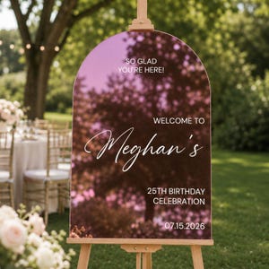 May include: An arched, rose gold mirrored acrylic welcome sign on a wooden easel. The sign features white text reading: SO GLAD YOU'RE HERE! WELCOME TO Meghan's 25TH BIRTHDAY CELEBRATION 07.15.2026. The surface reflects the surrounding outdoor trees.