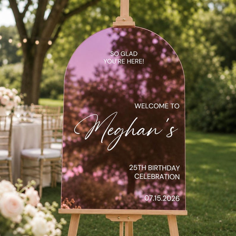 May include: An arched, rose gold mirrored acrylic welcome sign on a wooden easel. The sign features white text reading: SO GLAD YOU'RE HERE! WELCOME TO Meghan's 25TH BIRTHDAY CELEBRATION 07.15.2026. The surface reflects the surrounding outdoor trees.