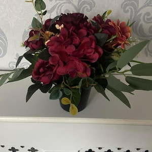 Può includere: Una composizione floreale con ortensie bordeaux scuro, rose e altri fiori con foglie verdi. I fiori sono in un vaso nero, creando un contrasto sorprendente. La composizione è posta su una superficie bianca.