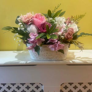 May include: A decorative floral display in a white metal basket. The arrangement features pink, white, and peach roses, complemented by green foliage and small white blossoms. The basket is inscribed with "FLOWERS & GARDEN".