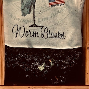May include: A white fabric worm blanket with the text "Worm Blanket" and "Made in USA" printed on it. The blanket is covering a wooden box filled with dark brown soil.