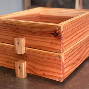 May include: Two-tiered wooden box with a natural reddish-brown finish. The box is constructed from planks of wood, with visible grain and screw fasteners. Small wooden blocks are attached to the sides for handles. The box is designed for storage or display.