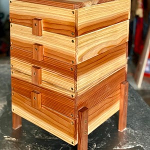 May include: A wooden beehive with a layered design, featuring natural wood tones and a boxy shape. The hive is supported by four wooden legs and secured with wooden pegs and screws. The overall dimensions are approximately 18 inches tall and 16 inches wide.