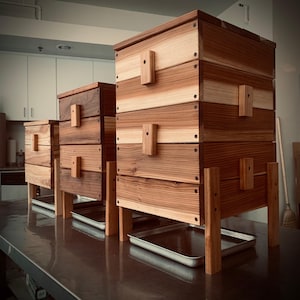 May include: Three wooden compost bins with lids and trays. The bins are made of natural wood with a light brown finish. The bins are stacked on top of each other, with the largest bin on the bottom and the smallest bin on top.