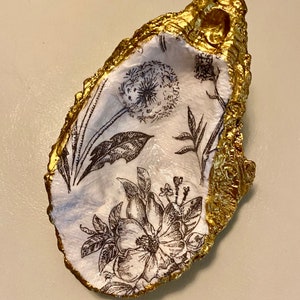 May include: A white oyster shell with a gold rim, decorated with a black and white floral pattern. The shell is a decorative dish or trinket holder.