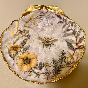 May include: A decorative seashell dish with a gold-painted rim. The interior features a bee and floral design in yellow, green, and white. The dish is a unique, handcrafted item.