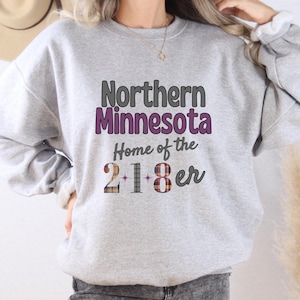 May include: A gray sweatshirt with the text "Northern Minnesota Home of the 21*8er" in purple and gray lettering. The numbers 2, 1, and 8 are in a plaid pattern.