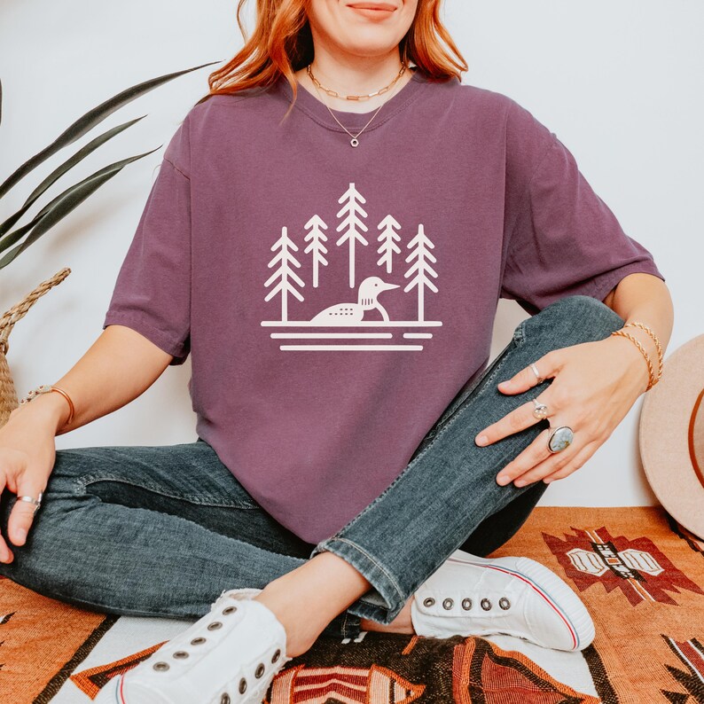 A relaxed-fit T-shirt in a rich berry color featuring a minimalist loon and pine tree design in white. The nature-inspired graphic showcases a loon on calm waters surrounded by tall evergreens. Styled with jeans, white sneakers, and boho jewelry, this shirt is perfect for outdoor lovers, lake enthusiasts, and those who appreciate cozy, nature-themed fashion.