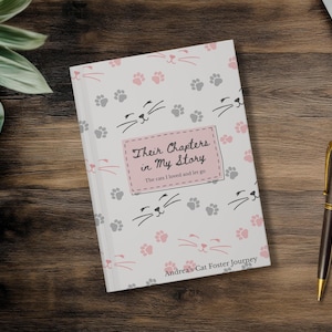 May include: A hardcover journal with a white cover featuring cat faces and paw prints in pink and gray. The title "Their Chapters in My Story" is displayed on a pink label. The journal is on a wooden surface.
