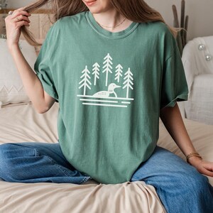 A relaxed-fit T-shirt in a soft light green color featuring a minimalist loon and pine tree design in white. The nature-inspired graphic showcases a loon floating on calm waters, surrounded by tall evergreens. Styled with casual blue jeans and delicate jewelry, this cozy and earthy tee is perfect for outdoor lovers, lake enthusiasts, and those who appreciate nature-inspired fashion.