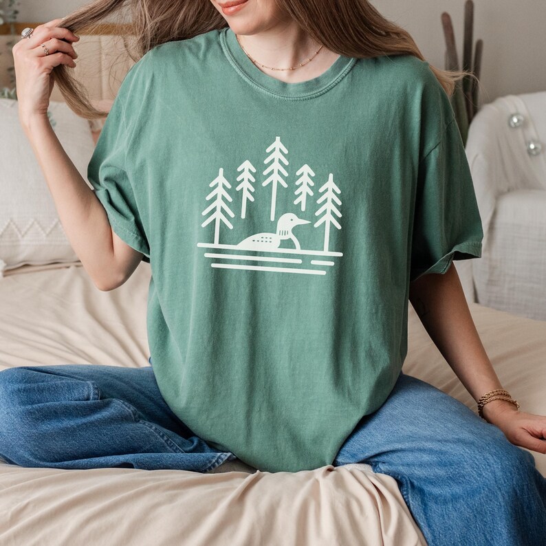 A relaxed-fit T-shirt in a soft light green color featuring a minimalist loon and pine tree design in white. The nature-inspired graphic showcases a loon floating on calm waters, surrounded by tall evergreens. Styled with casual blue jeans and delicate jewelry, this cozy and earthy tee is perfect for outdoor lovers, lake enthusiasts, and those who appreciate nature-inspired fashion.