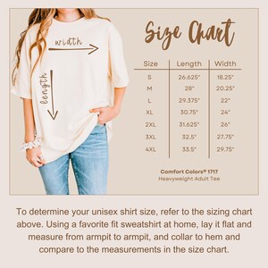 Comfort Colors® 1717 heavyweight adult tee size chart. The image features a model wearing an oversized cream-colored T-shirt with arrows indicating width and length. A detailed chart lists measurements for sizes S to 4XL. Below, text provides guidance on measuring a favorite sweatshirt at home for comparison. The neutral-toned design offers a clean and easy-to-read layout for size selection.