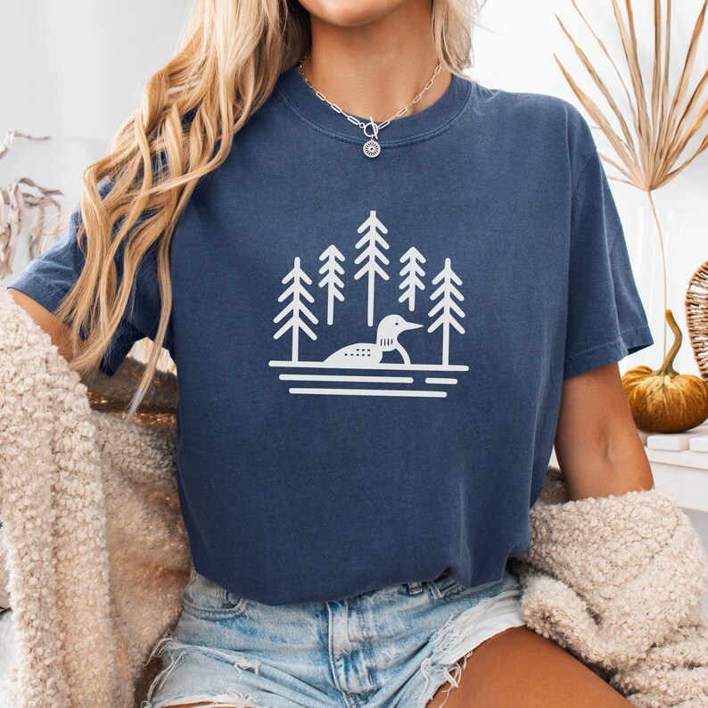 A relaxed-fit T-shirt in a deep denim blue color featuring a minimalist loon and pine tree design in white. The serene graphic showcases a loon gliding on calm waters, surrounded by tall evergreens. Styled with distressed denim shorts, a cozy knit cardigan, and a delicate silver necklace, this casual yet stylish tee is perfect for nature lovers, outdoor adventurers, and lake life enthusiasts.