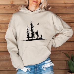 May include: A beige hoodie with a black silhouette of a person walking a dog through a forest with three trees. A bird flies overhead.