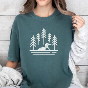 A cozy, relaxed-fit T-shirt in a muted blue spruce color featuring a minimalist loon and pine tree design in white. The graphic showcases a loon floating on calm waters, surrounded by tall evergreen trees, evoking a peaceful, nature-inspired aesthetic. Styled with a chunky knit cardigan and distressed jeans, this shirt is perfect for nature lovers, outdoor enthusiasts, and anyone who appreciates serene lake life.