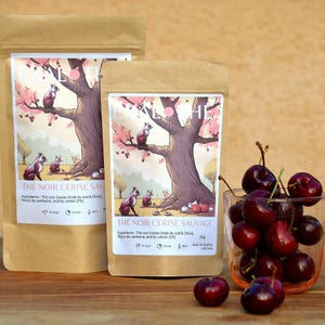 Könnte beinhalten: Zwei braune Papiertüten mit Illustrationen eines Baumes und Tieren. Die Tüten sind mit "The Noir Cerise Sauvage" beschriftet und enthalten 50g schwarzen Tee mit Kirschgeschmack. Die Tüten liegen auf einer Holzoberfläche mit Kirschen in einem Glas.