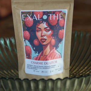 May include: A brown paper bag of tea with a colorful label featuring a woman with raspberries in her hair. The label reads "EXAL THE CHARME DU LITCHI". The bag is sitting in a clear glass bowl.