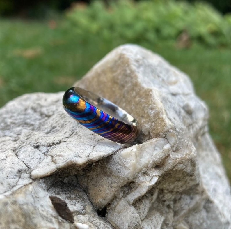 May include: A close-up of a colourful, iridescent ring with a silver band. The ring is made of a unique metal with a swirling pattern of blue, purple, and gold.