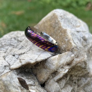 May include: A close-up of a colourful, patterned ring with a silver band. The ring is made of a metal with a unique, iridescent finish. The ring is resting on a grey rock.
